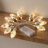 Warm white Christmas string lights with clear decorative bulbs and a connected translucent battery box rest on a wooden shelf.