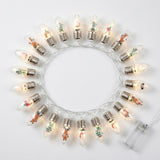 Christmas string lights with clear, silver-capped bulbs and miniature festive figures glow in a circle on a white background beside a battery box.