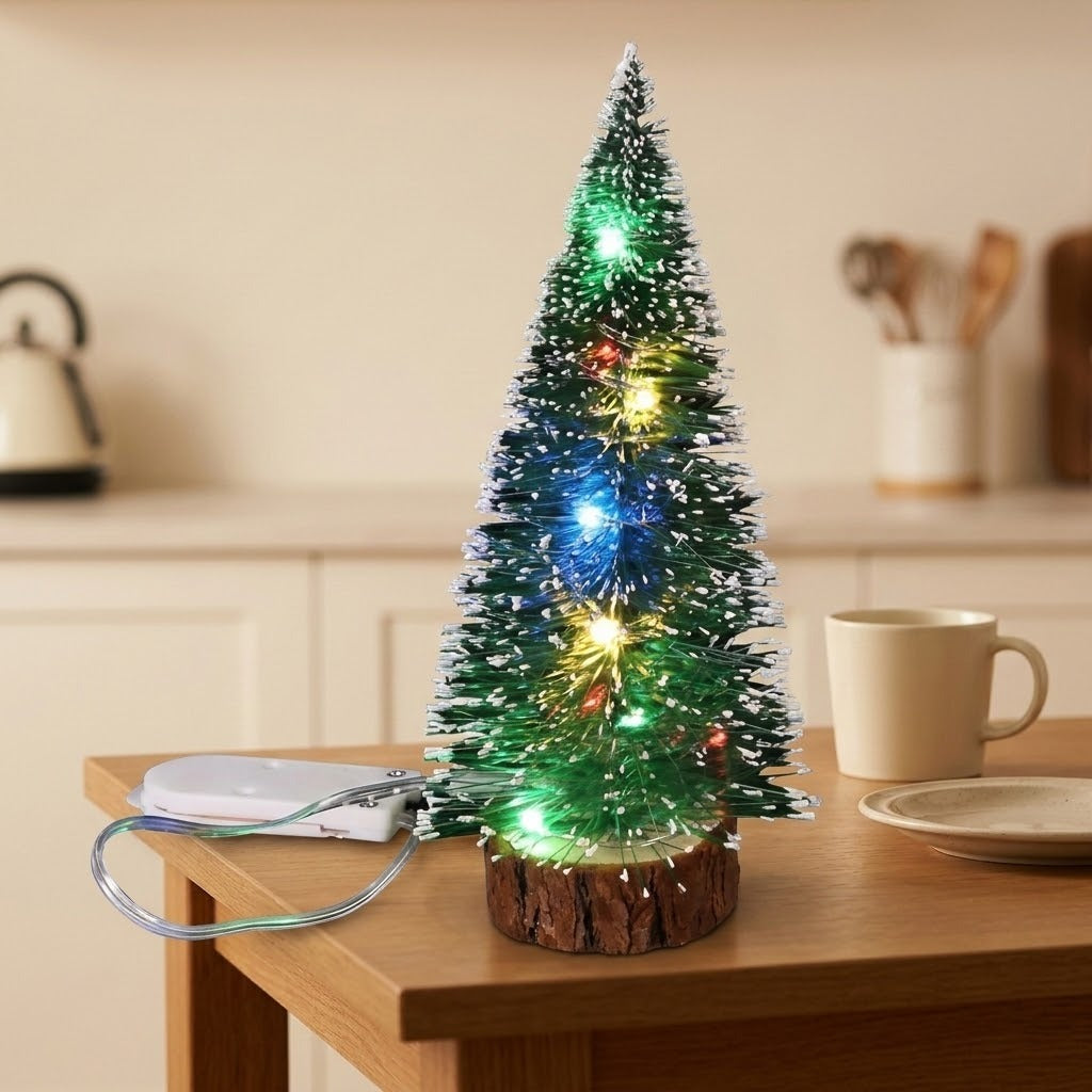 A snow-frosted green mini Christmas tree with multicolor LED lights and a white battery pack stands on a kitchen table beside a mug.