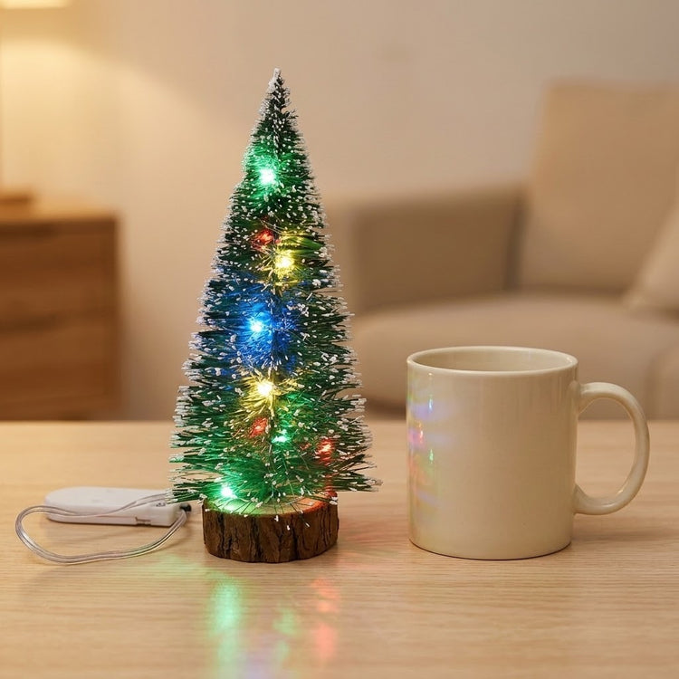 A snow-frosted mini Christmas tree with multicolor LED lights and a wooden-look base sits beside a cream mug, with a sofa behind.