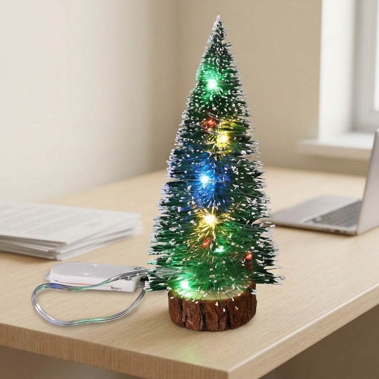 A snow-frosted mini Christmas tree with multicolor LED lights and an attached white battery pack sits on a desk beside a laptop.