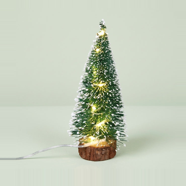 A snow-frosted green mini Christmas tree with warm white lights stands on a bark-edged wood base against a pale green background.