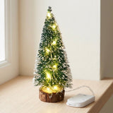 A snow-frosted green mini Christmas tree with warm white lights stands on a wooden windowsill beside a white battery pack.