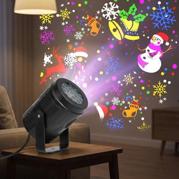 A black holiday light projector on a wooden side table casts colorful snowflakes, bells, stockings, a reindeer, and a snowman onto the wall.