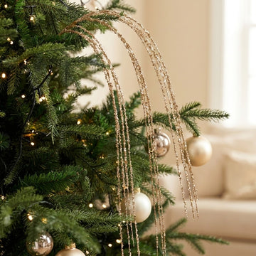 A gold glitter Christmas tree pick cascades over green branches beside white and metallic baubles, with a bright window behind.