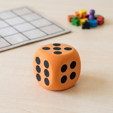 An orange foam die with black dots sits on a light wood tabletop beside a blurred game board and colorful playing pieces.