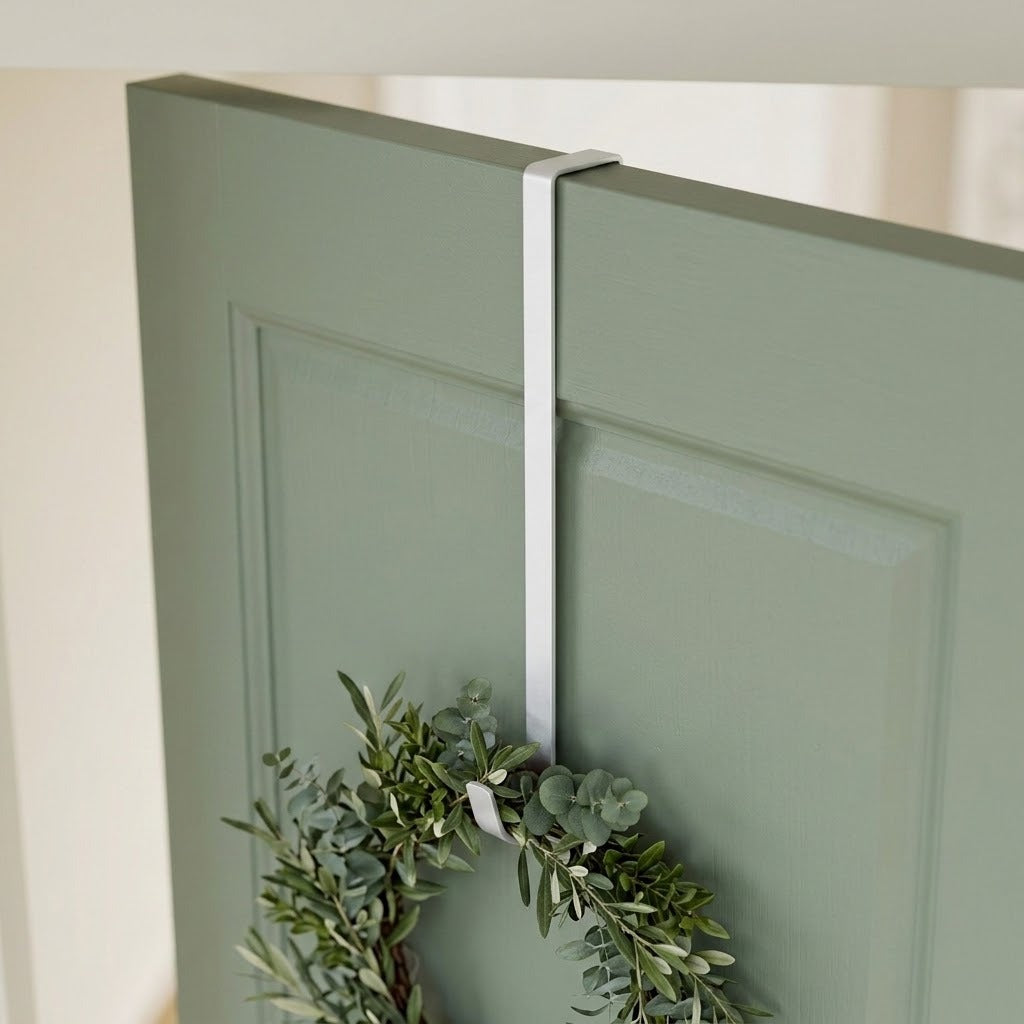 A white zinc alloy wreath hanger fits over a sage green paneled door, supporting a leafy green wreath.