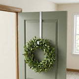 A white wreath hanger holds a leafy wreath with green berries on an open sage green door in a bright entryway.