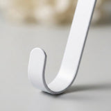 A close-up shows the white zinc alloy wreath hanger's rounded lower hook resting on a gray surface against a blurred neutral background.