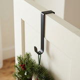 A black wreath hanger hooks over a white door, supporting evergreen foliage and red berries from a narrow green loop.