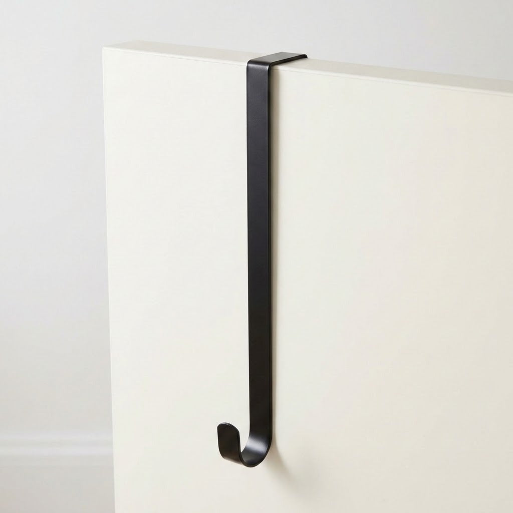 A matte black wreath hanger rests over a white door, showing its long flat shaft and curved lower hook.
