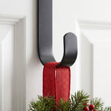 A black wreath hanger holds a red ribbon supporting evergreen foliage and red berries against a white paneled door.