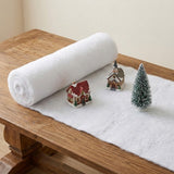 A white snow blanket sits partially unrolled on a wooden table beneath miniature buildings and a snow-dusted green tree.