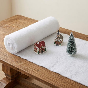 A white snow blanket sits partially unrolled on a wooden table beneath miniature buildings and a snow-dusted green tree.