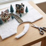 A white snow blanket with a curved cutout supports miniature houses, trees, and a bridge on a wooden table beside scissors.