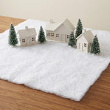 A fluffy white snow blanket covers a wooden tabletop beneath three miniature cream houses and snow-dusted green trees.