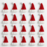 Miniature Christmas accessories include twenty red knitted Santa hats with white cuffs and pom-poms arranged in four rows on white.