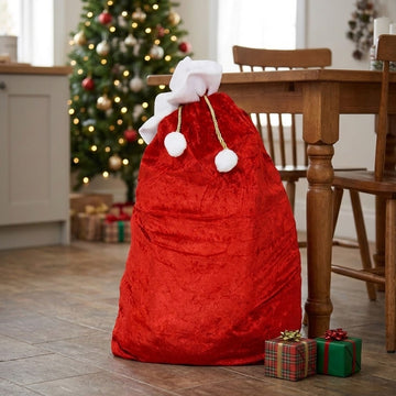 A large red Christmas Santa sack with white faux fur trim and drawstring cords sits on a wooden floor in a dining room, with a decorated Christmas tree, wooden table, and small wrapped gifts in the background.
