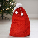 A red fleece gift sack with white trim and pompoms stands on a pale floor beside a decorated Christmas tree.