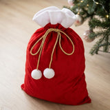 A red fleece gift sack tied with a gold bow stands on pale wood beside a Christmas tree, with white trim and pompoms.