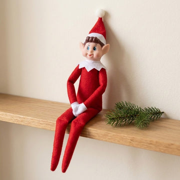 A red elf doll with white gloves and collar sits on a wooden shelf beside an evergreen sprig against a cream wall.