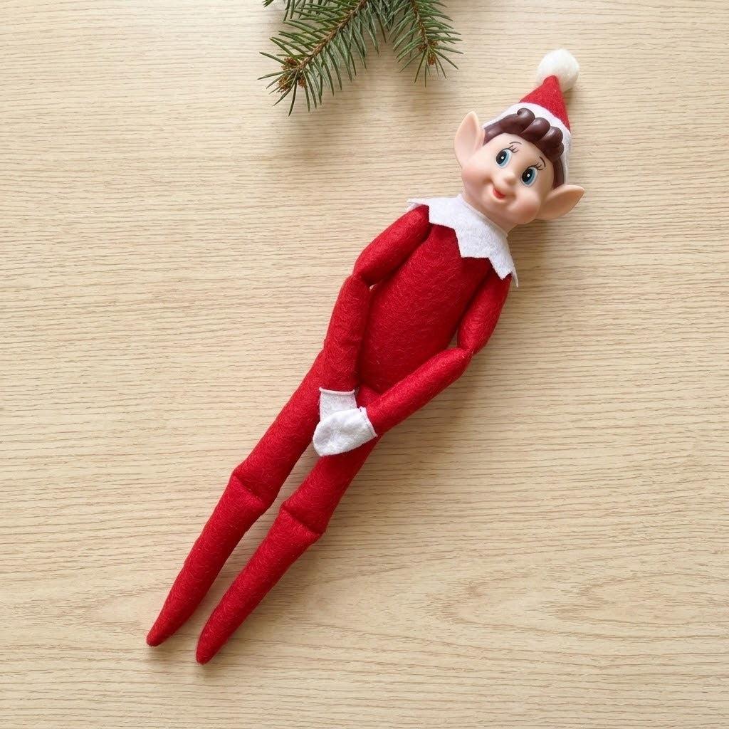 A red elf doll with brown hair and white trim lies diagonally on a light wooden surface beneath an evergreen sprig.