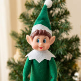 A close-up of a green elf doll shows brown molded hair, blue eyes, pointed ears, and a white collar before a Christmas tree.