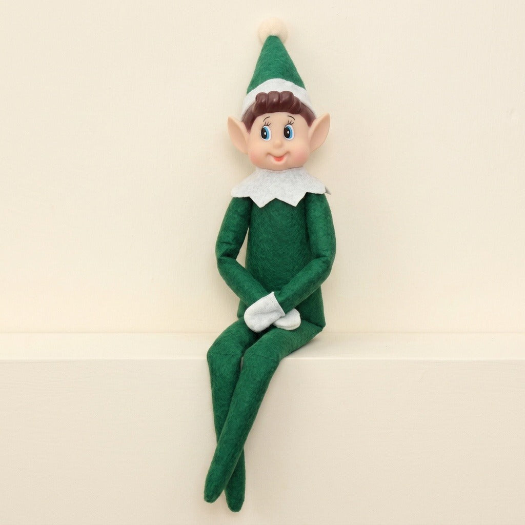 A green elf doll with white gloves and collar sits with crossed legs on a cream ledge against a matching wall.