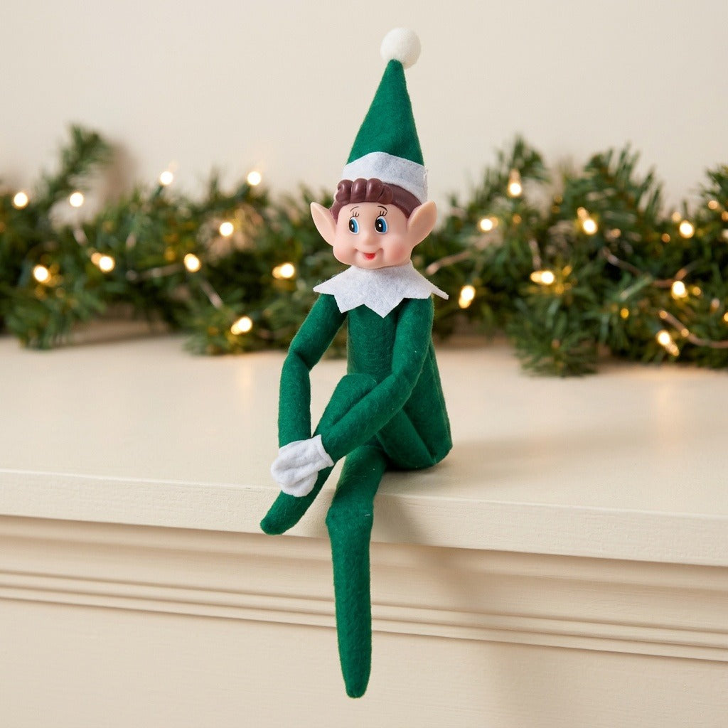 A green elf doll sits with one leg crossed over the other on a cream mantel before evergreen garland and warm lights.