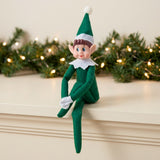 A green elf doll sits with one leg crossed over the other on a cream mantel before evergreen garland and warm lights.