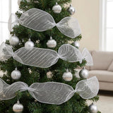 Silver mesh decorative ribbon forms wide gathered loops around a green Christmas tree with silver baubles beside a beige sofa.