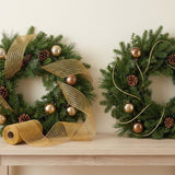 Gold mesh decorative ribbon wraps around an evergreen wreath beside a second wreath, with a loose roll on the wooden console.