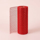 Red mesh decorative ribbon stands in an upright roll with a loose section extending left against a pale pink background.