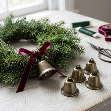 Bronze-colored iron craft bells sit beside a pine wreath, with one tied beneath a burgundy bow on a white wooden table.