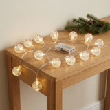Warm white string lights with clear bubble globes spread across a wooden console beside evergreen branches and a transparent battery box.