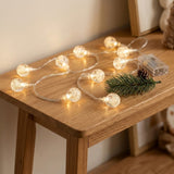Warm yellow globe string lights lie on a wooden table beside an evergreen sprig, pinecone, and clear battery box.