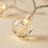 A close-up of string lights shows a clear bubble globe glowing warm yellow on beige fabric, attached to twisted silver-colored wire.