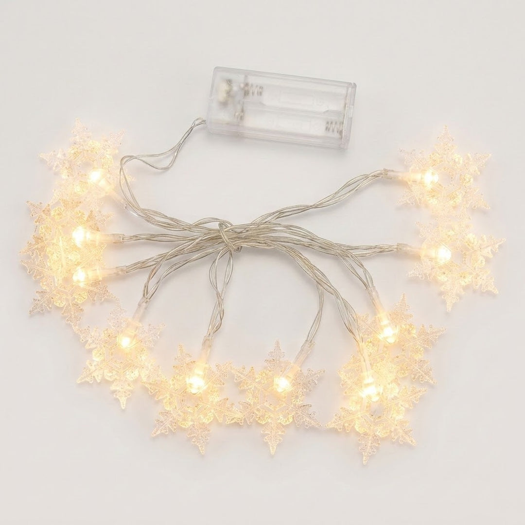 Clear snowflake string lights glow warm white around bundled silver-colored wires beneath a transparent battery box on a white background.