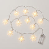 Clear star and snowflake string lights glow warm white along looping silver-colored wires beside a transparent battery box on white.