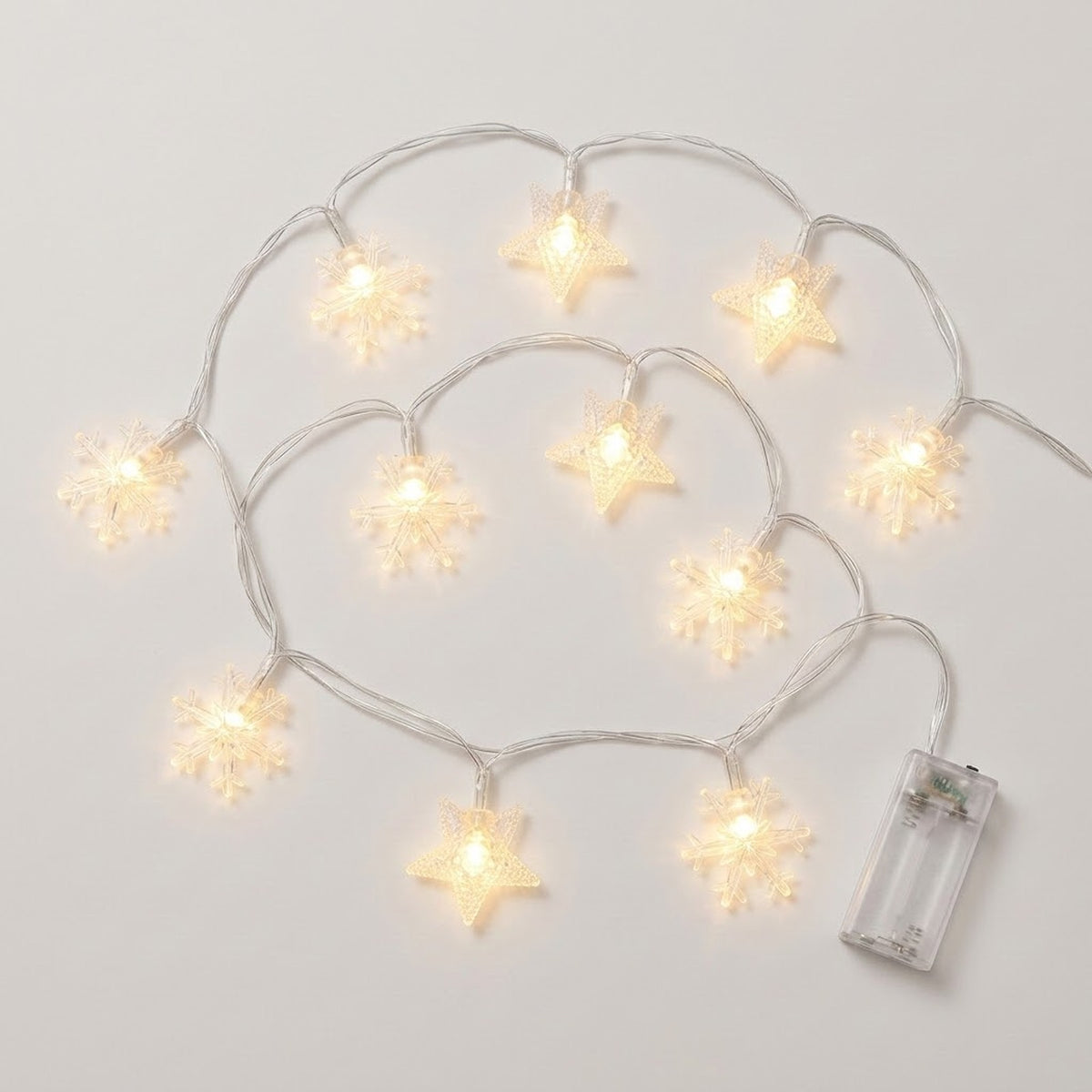 Clear star and snowflake string lights glow warm white along looping silver-colored wires beside a transparent battery box on white.