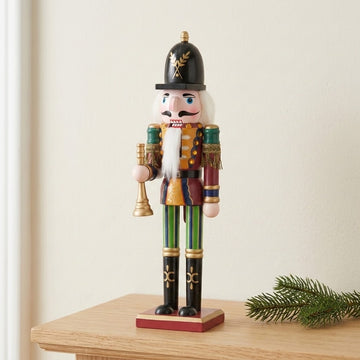 A burgundy and gold decorative nutcracker holds a gold trumpet on a wooden table beside an evergreen sprig against a cream wall.
