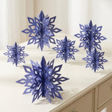Six dark blue 3D snowflake ornaments in assorted sizes stand on a cream console table beside white curtains.