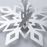 A silver snowflake ornament close-up shows reflective surfaces, diamond-shaped cutouts, and intersecting central folds against a pale background.
