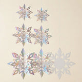 Iridescent silver snowflake ornaments hang against a cream background, showing six folded 3D shapes beside one flat snowflake.