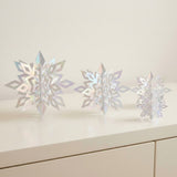 Three iridescent silver 3D snowflake ornaments stand in descending sizes on a white cabinet against a cream wall.