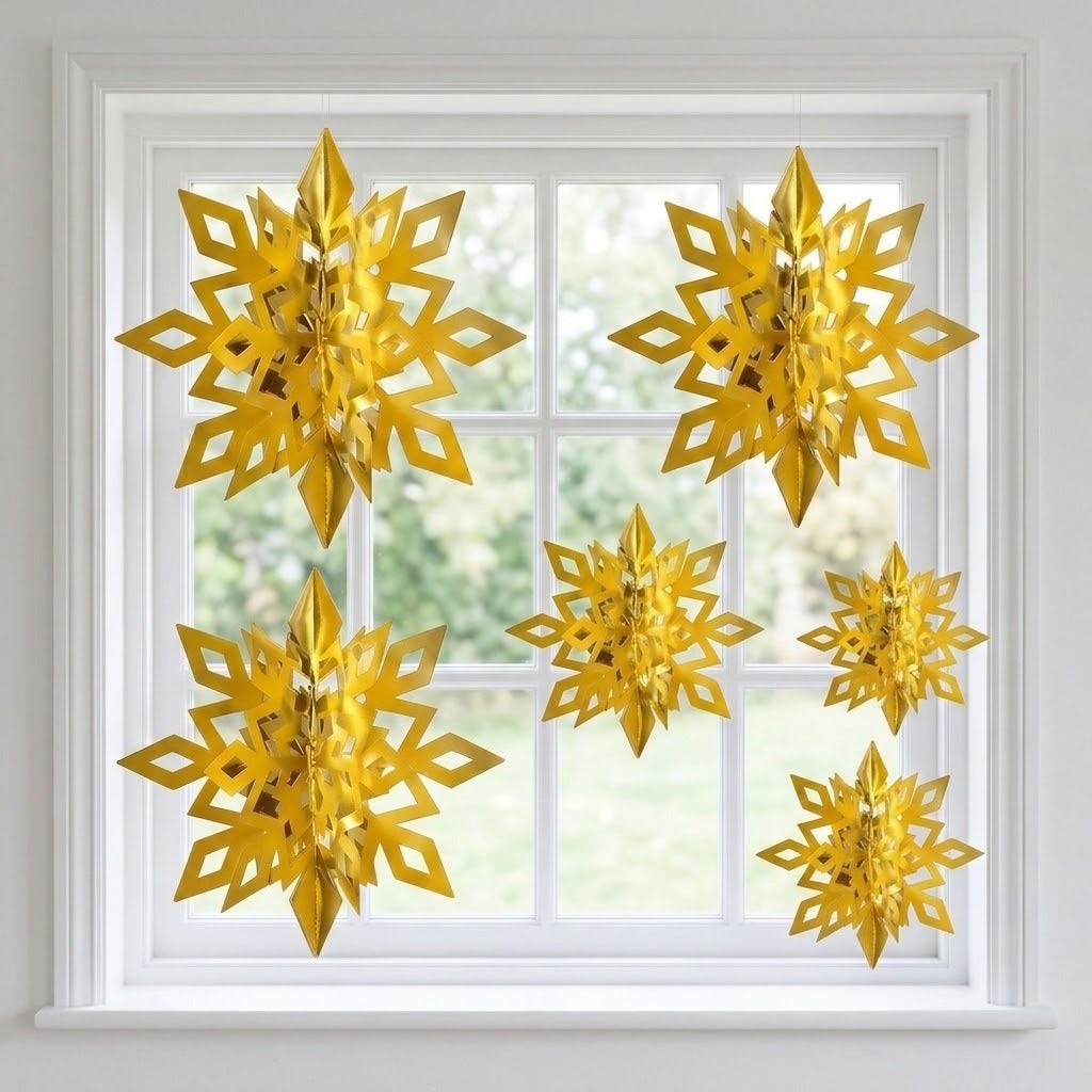 Six reflective gold 3D snowflake ornaments in assorted sizes hang across a white-framed window with blurred greenery outside.