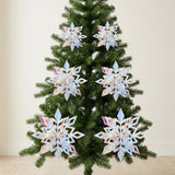 Six iridescent silver 3D snowflake ornaments hang in pairs on a green Christmas tree against a cream wall.