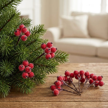 Frosted red artificial berry picks decorate green tree branches and rest on a rustic wooden table, with a blurred cream sofa behind.