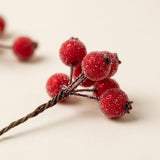 An artificial berry pick shows textured red berries and a twisted brown stem in close-up against a warm off-white background.