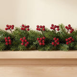 Red artificial berry picks are tucked into a green pine garland on a light wooden mantel against a cream wall.